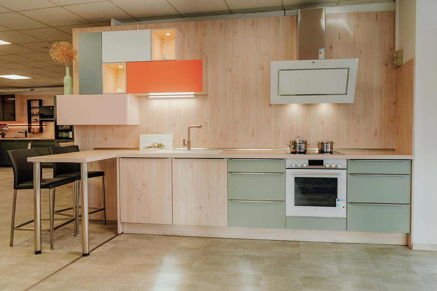Küche aus Holz mit Fronten in orange & mintgrün und weißer Dunstabzugshaube und Backofen in der Küchenausstellung Matthäs